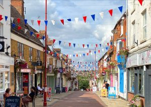 A view of Sheaf Street in Daventry decorated with red, white and blue bunting, with independent shops and people sitting outside cafés.