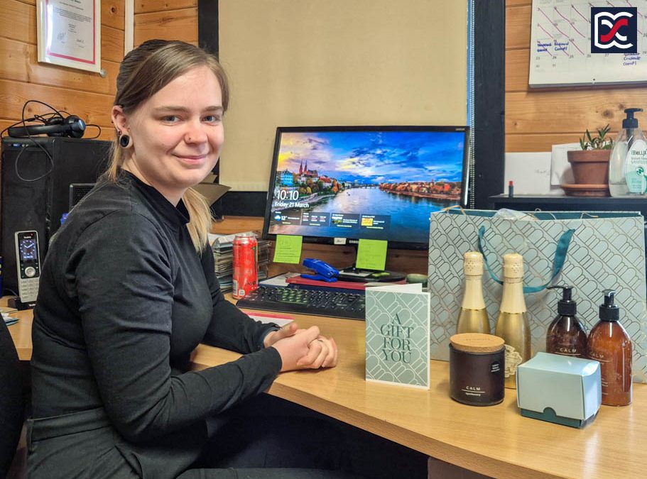 A team member sitting at a desk in an office, smiling beside a thank-you gift set from clients.