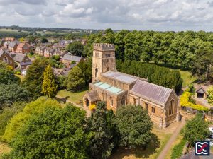 Aerial view of All Saints Church in Earls Barton, showing the distinctive Saxon tower surrounded by trees and village housing.